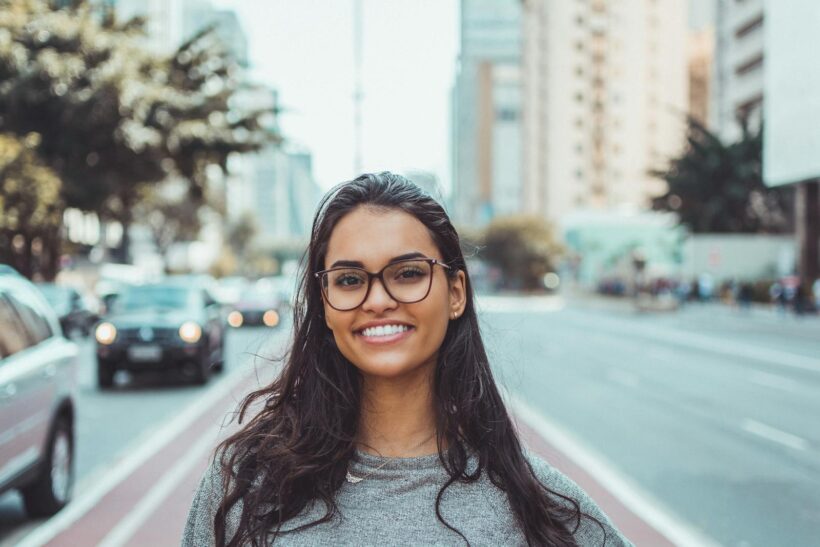 A young woman with glasses smiling on a city street, embracing urban lifestyle.